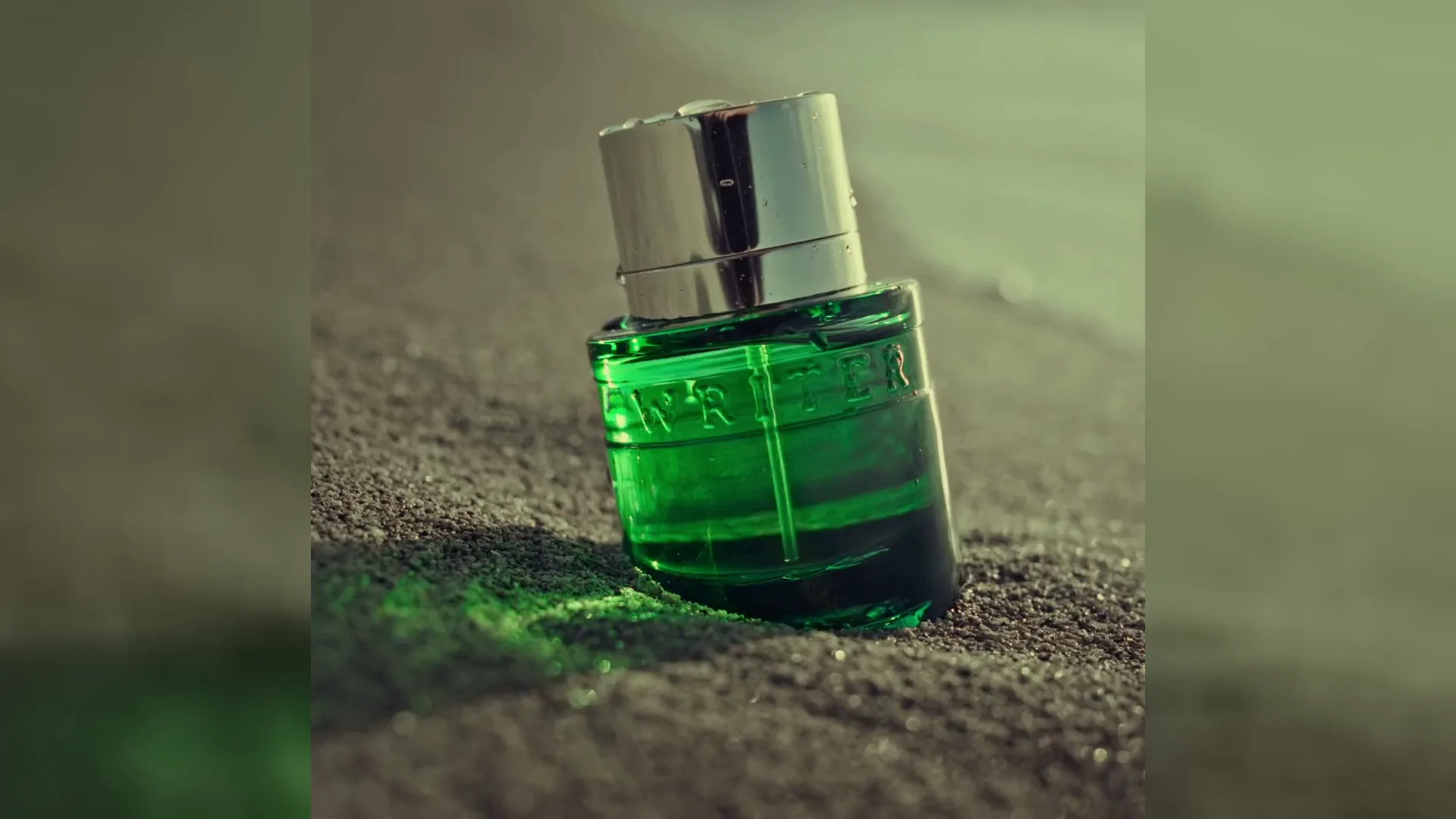 A green glass perfume bottle rests in sand at the water’s edge at sunset.
